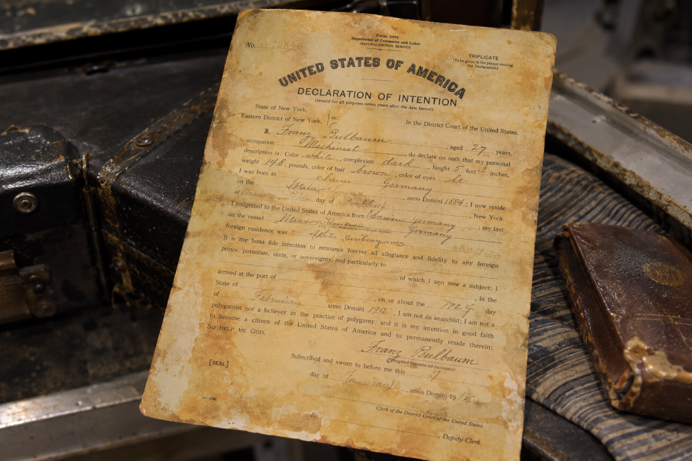 A vintage U.S. Declaration of Intention form for Franz Baulbaum, dated 1912, resting on a worn suitcase next to a small book.