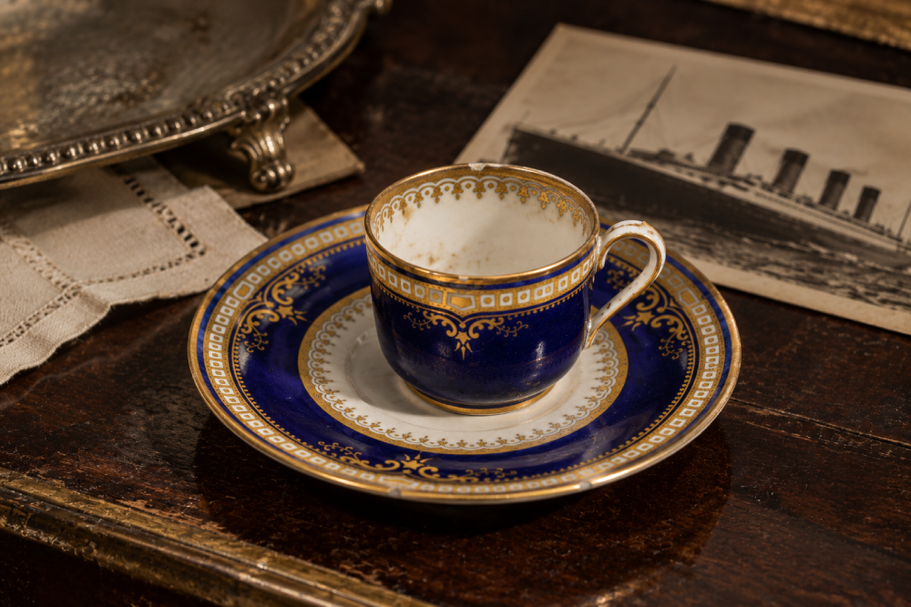 A vintage navy blue and gold teacup and saucer set resting on a dark wooden surface next to an old postcard of a ship.