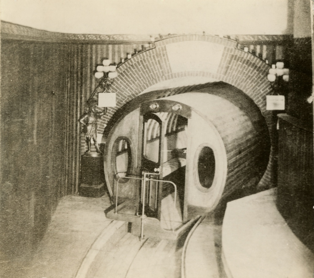 A rounded, barrel-shaped subway car sits in an arched tunnel station, with a decorative statue standing on a pedestal nearby.