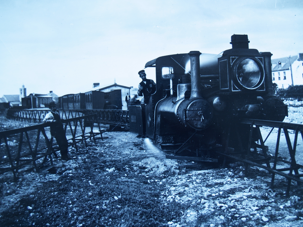 A steam locomotive on a rail line with a train conductor in the cab and a worker standing by the tracks.