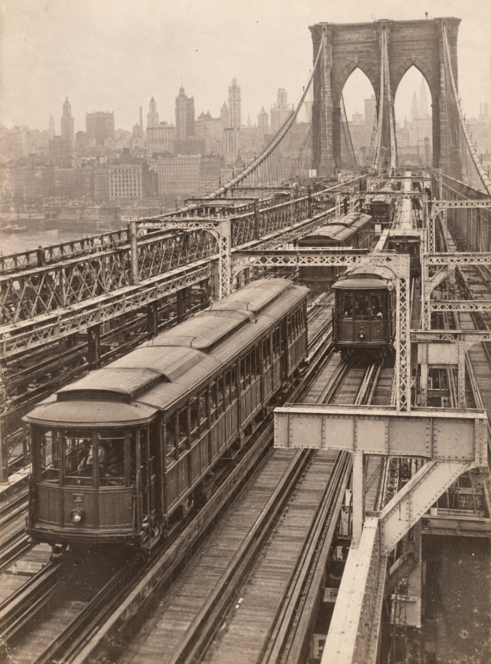 Two vintage trolley cars travel along the tracks of the Brooklyn Bridge with the New York City skyline in the background.
