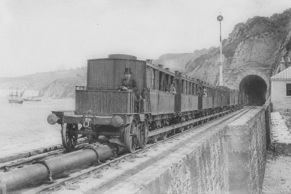 A train with a man in a top hat at the front travels along a seaside track leading into a tunnel, with a large pipe running between the rails.