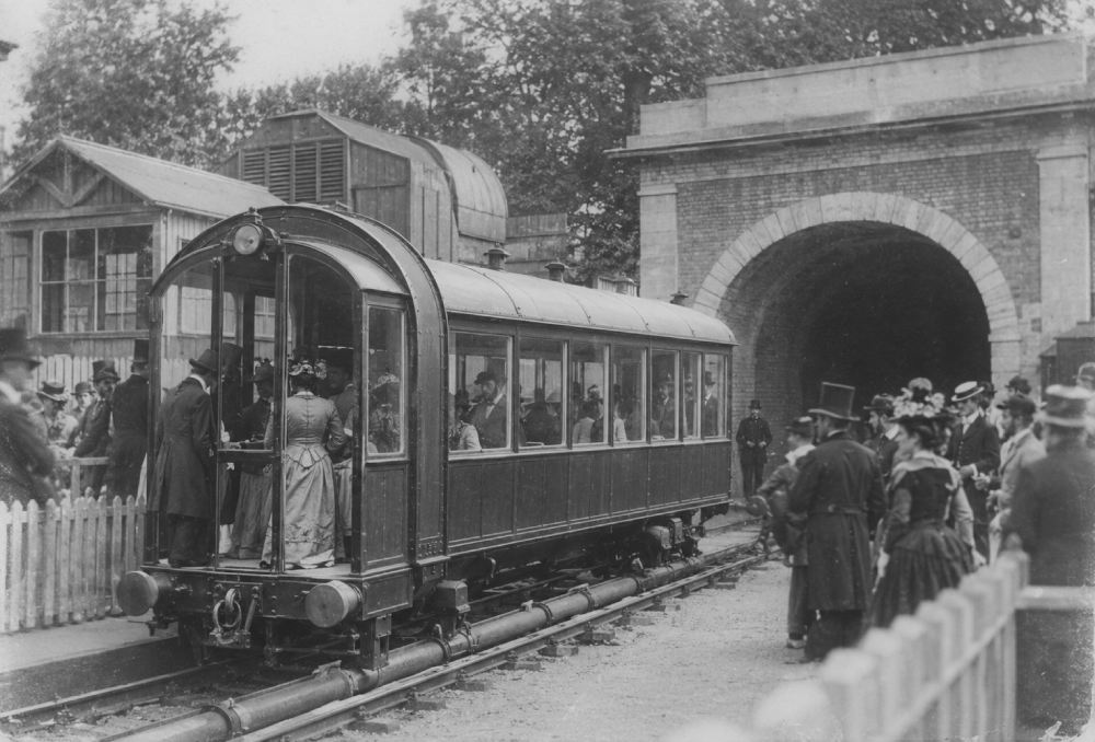 A group of Victorian-era passengers in formal clothing board a rail car near the entrance to a brick tunnel.