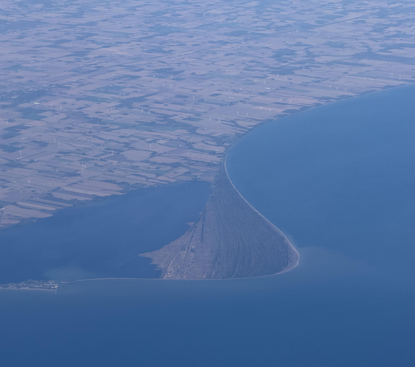 Aerial view of a long, forested sand spit extending into a large body of water, with a grid of agricultural fields and wind turbines on the adjacent coastline.