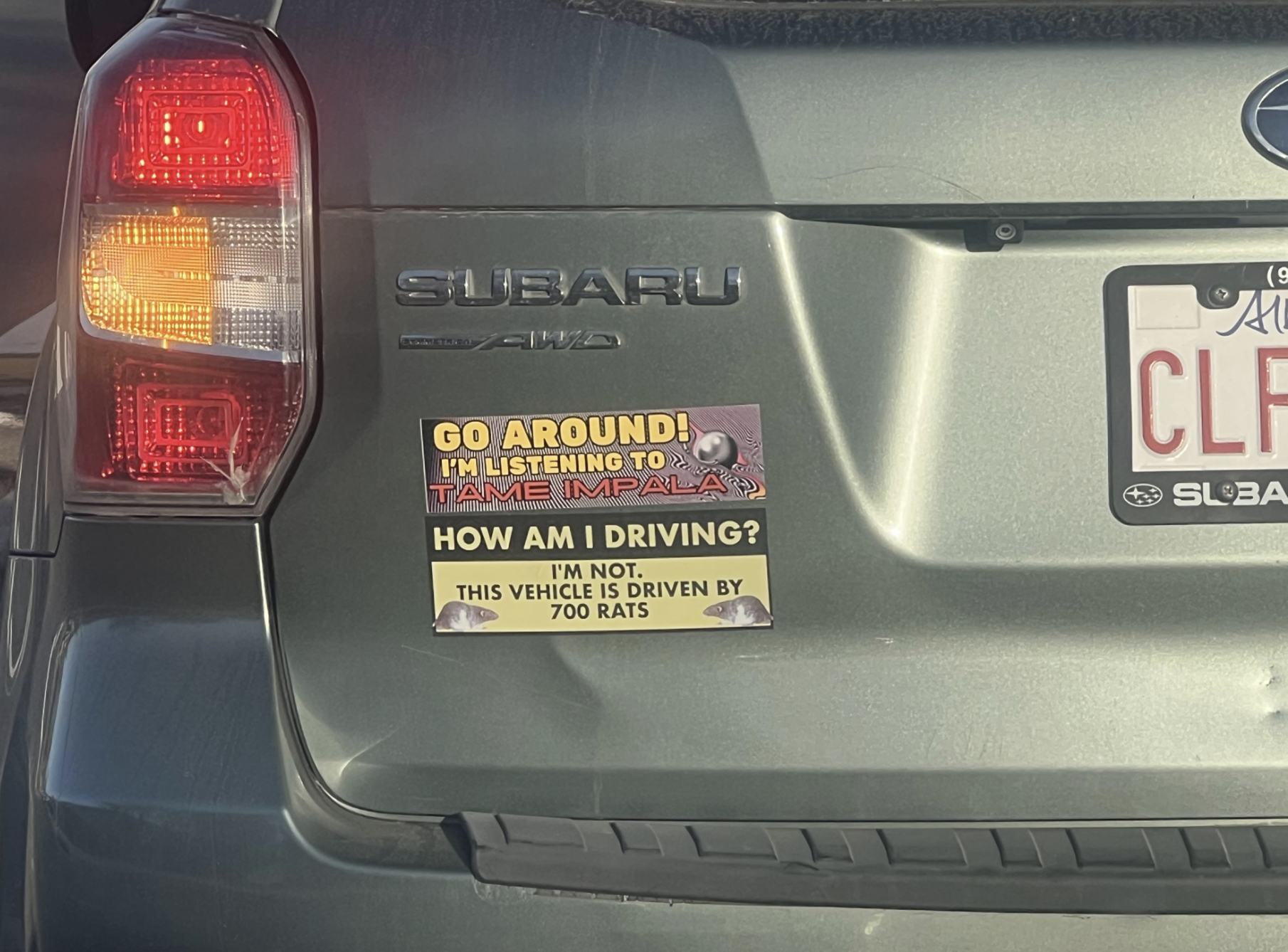 Two bumper stickers on a gray Subaru: the top reads “GO AROUND! I’M LISTENING TO TAME IMPALA,” and the bottom reads “HOW AM I DRIVING? I’M NOT. THIS VEHICLE IS DRIVEN BY 700 RATS.”