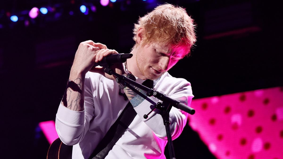 A musician with reddish hair in a white shirt holds a microphone and guitar on stage under purple and pink lights.