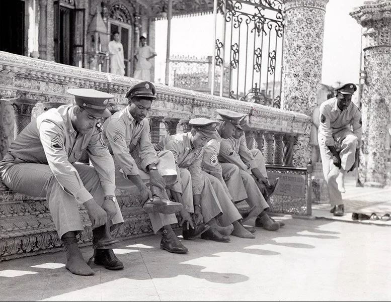 Five U.S. soldiers in uniform sit on a stone ledge outside an ornate building, putting on their boots, while a sixth soldier stands nearby adjusting his own footwear.