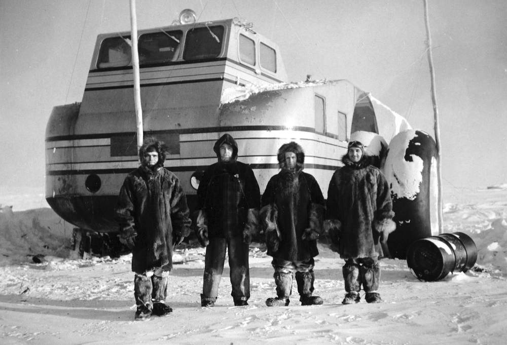 Four people in heavy fur-lined winter gear stand in front of a large Antarctic snow cruiser.