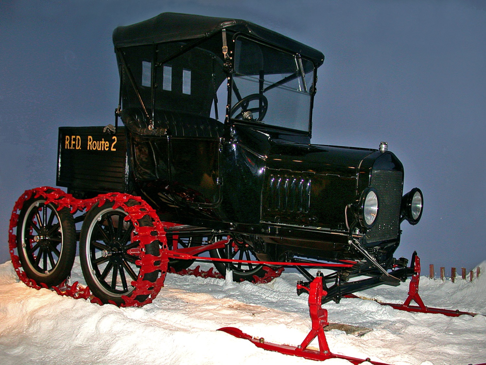 A vintage black Ford Model T converted for snow travel, with red tracks on the rear wheels and red skis mounted to the front wheels, labeled “R.F.D. Route 2” on the side.