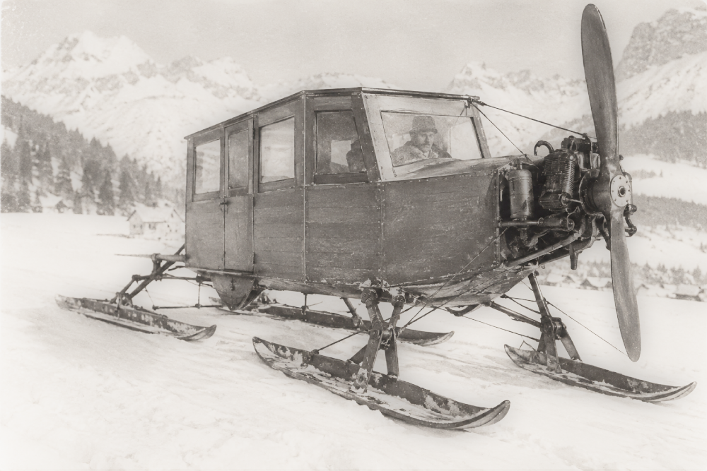 A vintage propeller-driven snow vehicle with an enclosed wooden cabin mounted on three skis, parked in a snowy mountain landscape.