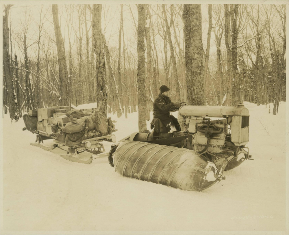 A person operates a snow vehicle with large, ribbed metal rollers instead of wheels, pulling a sled loaded with supplies through a snowy forest.