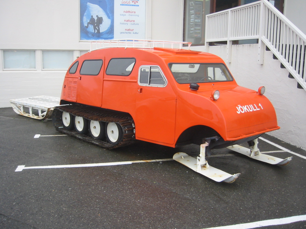 Bright orange snowcat vehicle with front skis and rear tracks, labeled “JÖKULL 1,” parked in a lot.