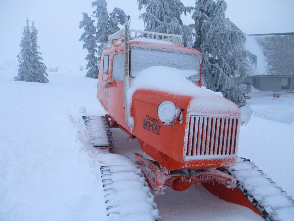 An orange Tucker Sno-Cat parked in deep snow in front of frost-covered trees and a building.
