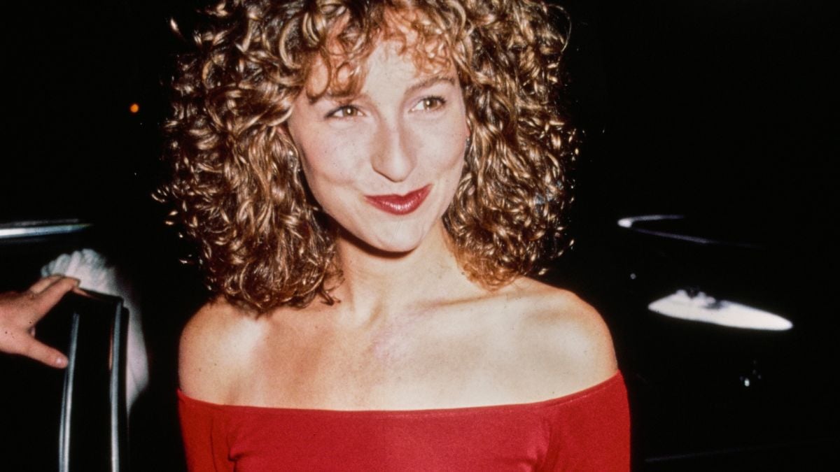 A woman with curly hair wearing an off-the-shoulder red top smiles, standing in a dimly lit setting with dark background and some lights.