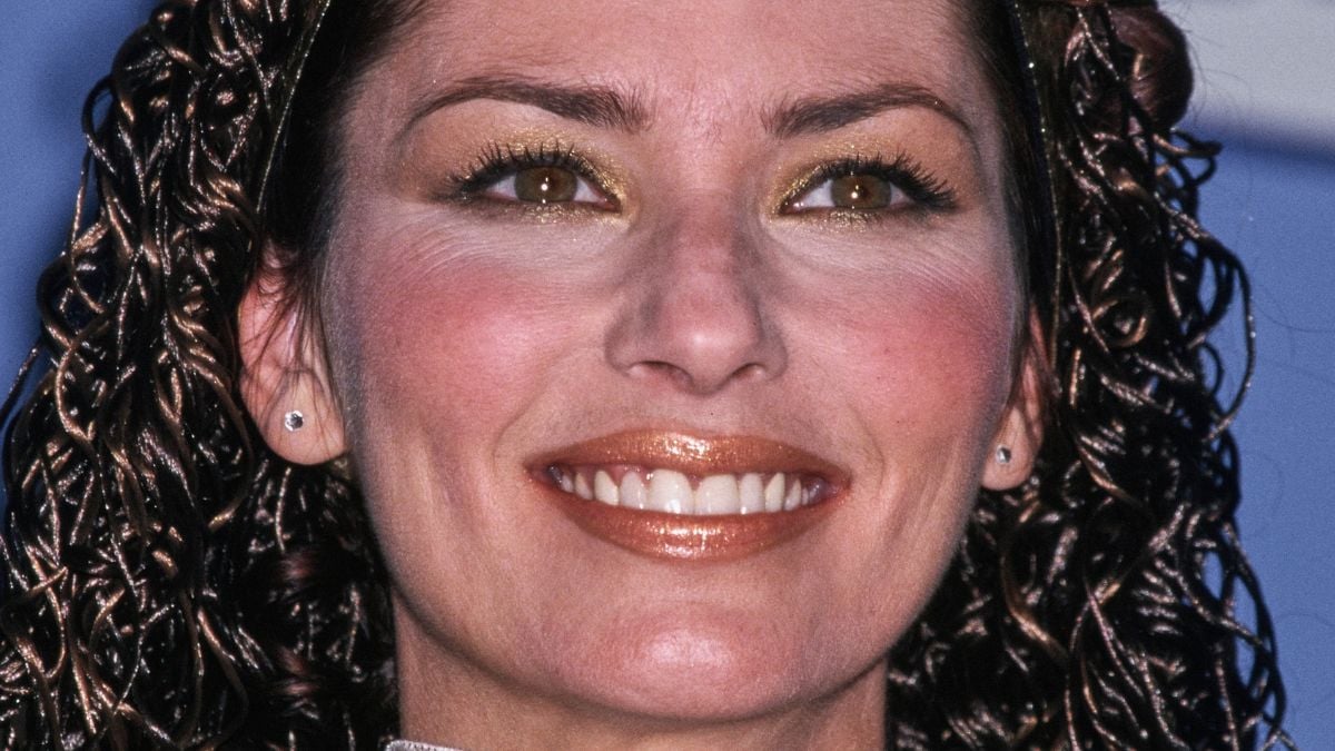 A woman with brown curly hair, golden eye makeup, and glossy lipstick smiles at the camera. She is wearing small stud earrings.
