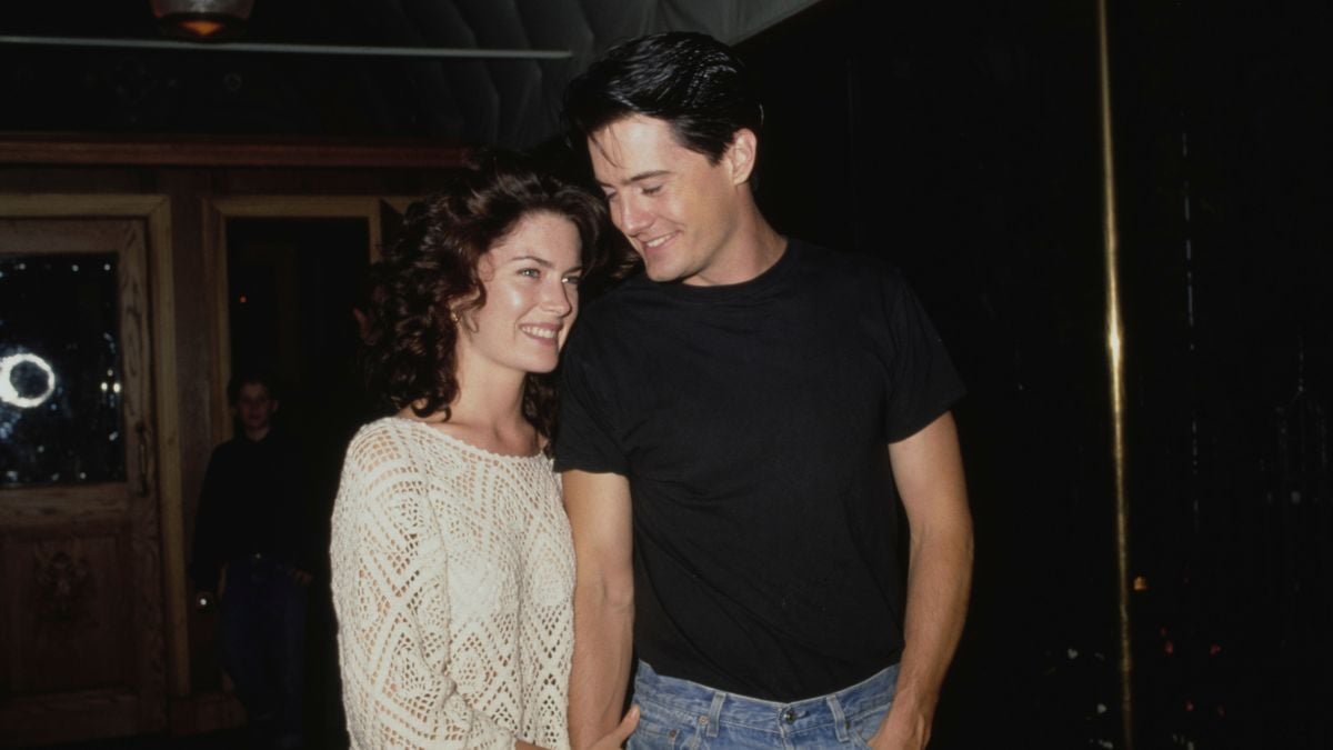 A woman in a white sweater smiles at a man in a black t-shirt and blue jeans. They stand close together indoors, looking happy and relaxed.
