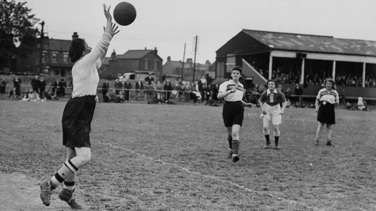 A vintage black-and-white photo of women playing soccer; one player throws a ball while others run toward her on a grassy field, with spectators and buildings in the background.