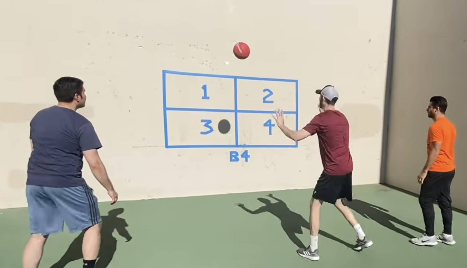 Three men play a wall ball game against a target grid marked one through four painted on a court wall.