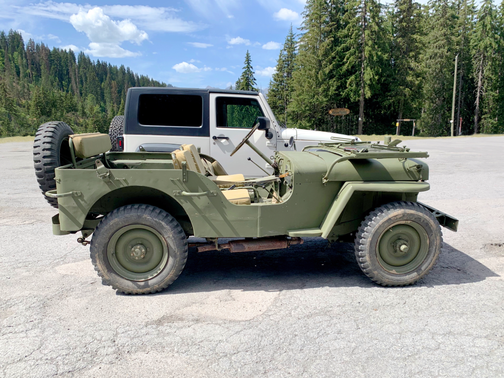 A vintage olive green military-style Jeep parked in a lot in front of a white modern Jeep SUV.