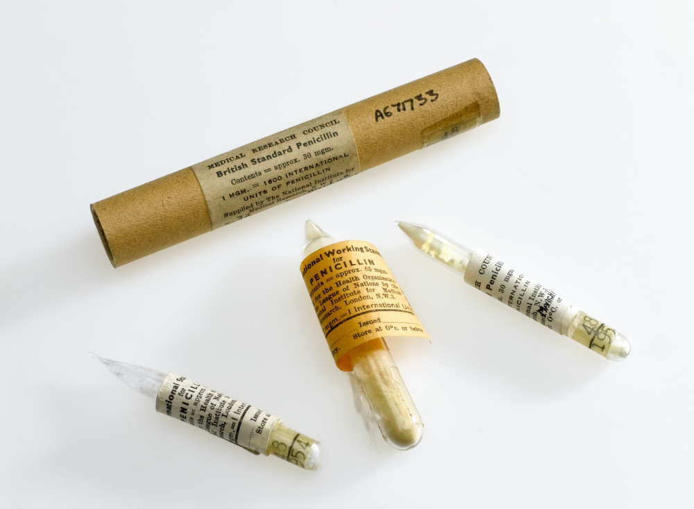 A cylindrical cardboard container and three glass ampoules, each wrapped in a label detailing British Standard Penicillin from the Medical Research Council.