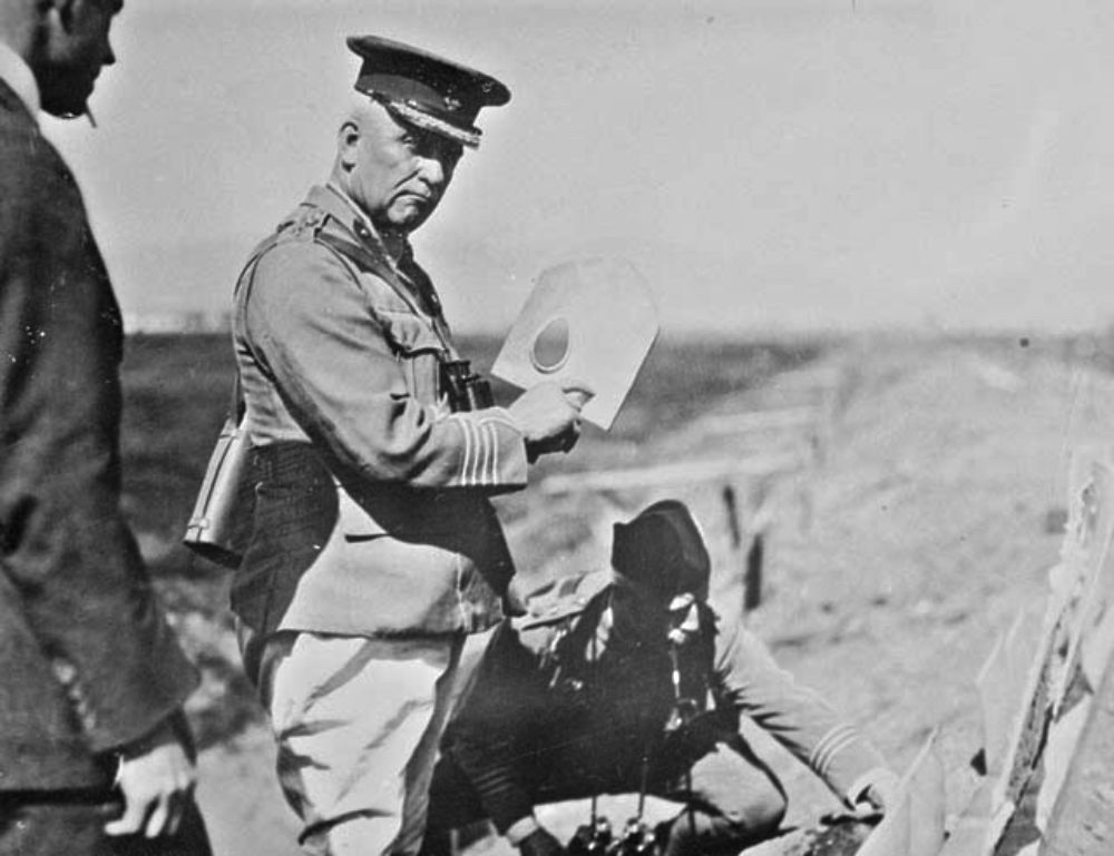 A military officer in uniform holds up a white board with a circular cutout as another soldier kneels on the ground nearby.