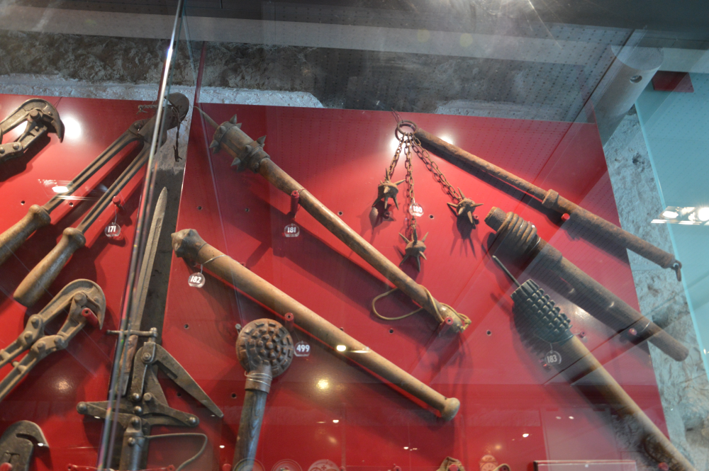 A display of various antique medieval flails, maces, and clubs mounted on a red wall inside a glass case.