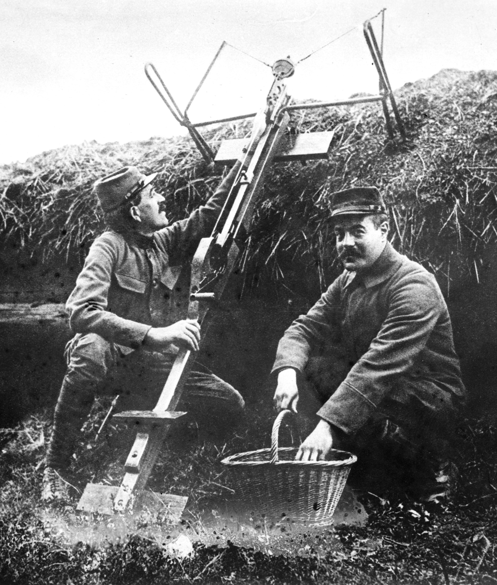 Two French soldiers in a trench operating a handheld mortar, with one holding a basket of grenades.