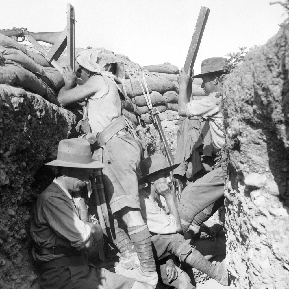 Soldiers in slouch hats stand and sit inside a dirt trench lined with sandbags.