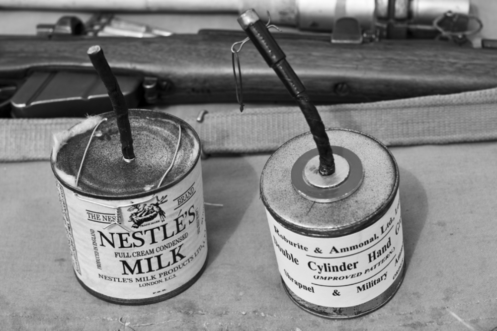 Two improvised hand grenades made from metal tins, one labeled as Nestlé’s Milk and the other as Roburite and Ammonal, sitting in front of a rifle.
