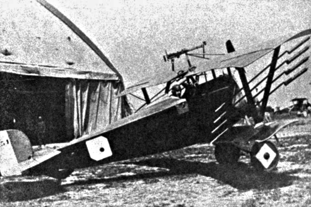 World War I-era biplane with a mounted machine gun, parked on an airfield near a hangar.