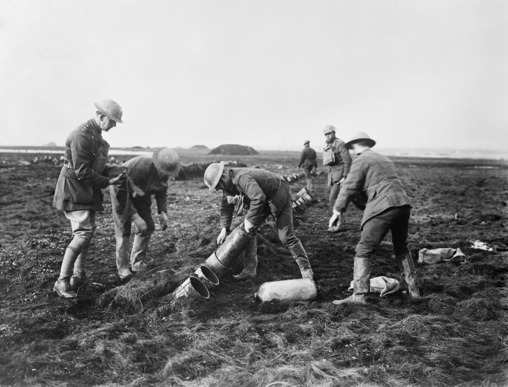 Soldiers in uniform and helmets placing metal canisters into the ground in a field.