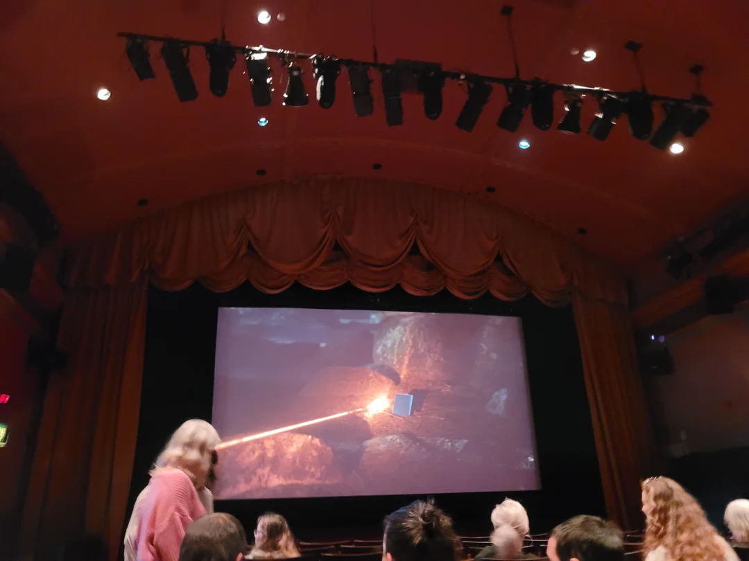 A person in a theater auditorium uses a laser pointer on a large screen displaying a rocky scene.