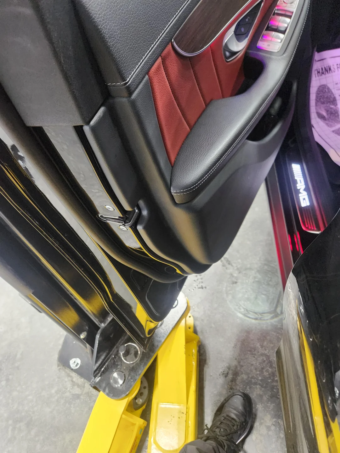 Open door of a vehicle with red and black interior panels and an illuminated AMG door sill, positioned over a yellow automotive lift arm.