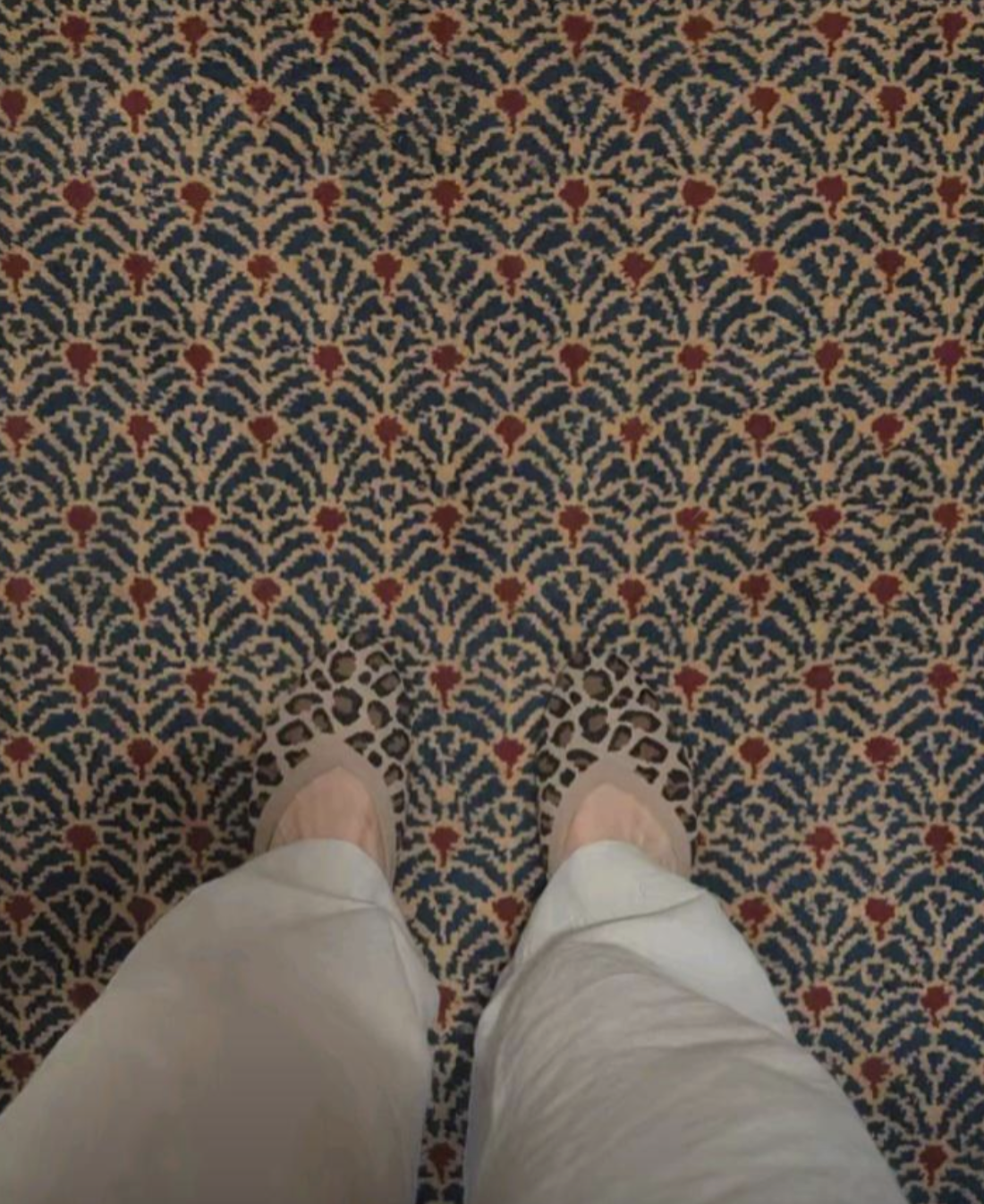 A person wearing light-colored trousers and leopard-print flats stands on a blue, red, and beige patterned carpet.