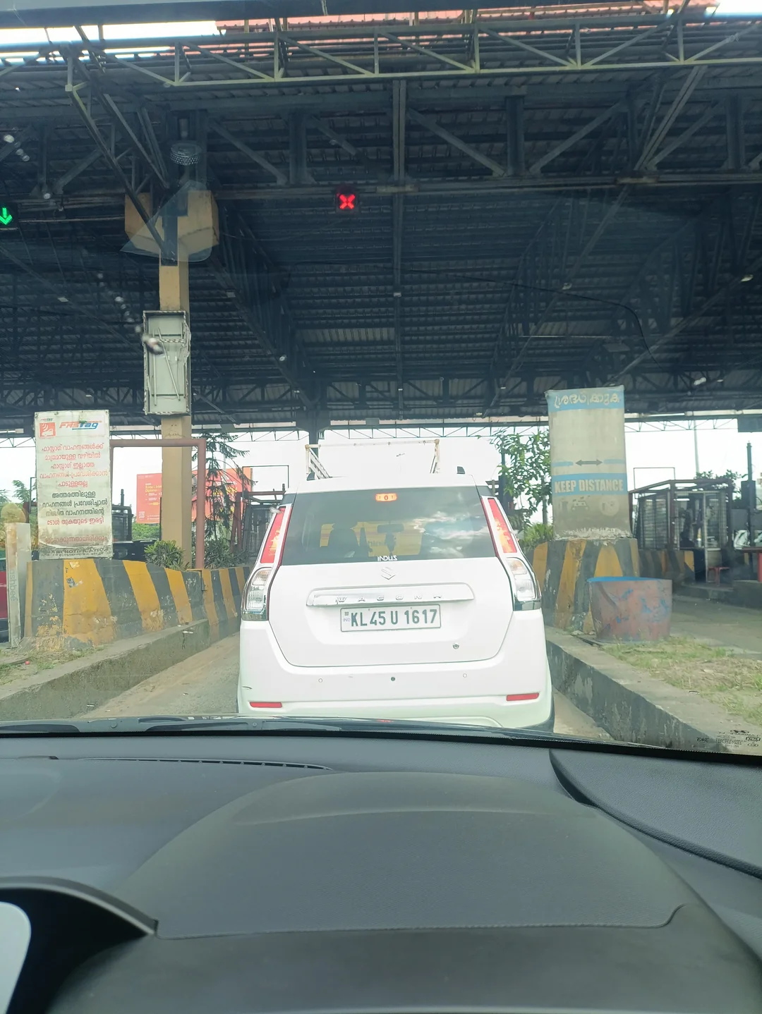 A white hatchback car with license plate KL 45 U 1617 waiting at a toll booth under a large metal canopy.