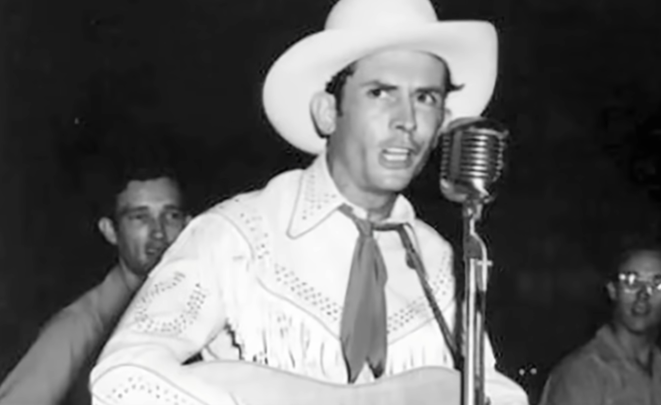 Hank Williams wearing a cowboy hat and fringe shirt singing into a vintage microphone.
