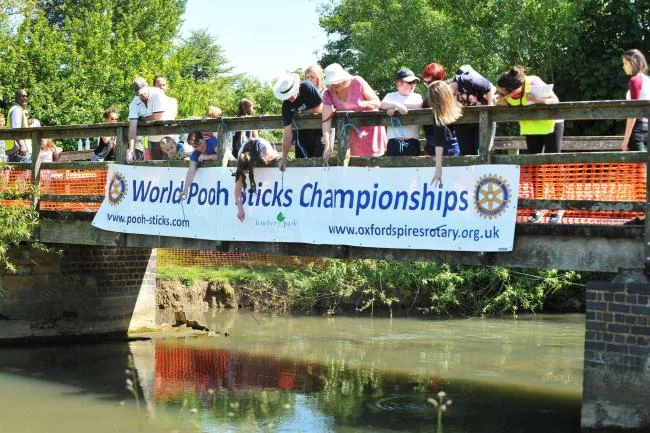 People lean over a bridge railing to watch the World Pooh Sticks Championships, marked by a large banner attached to the side of the bridge.